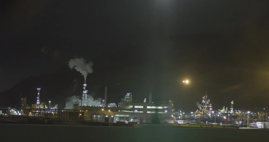 Smoke rises from a tall tower at a refinery in the middle of a city light up at night
