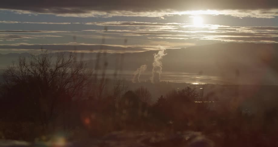 Steam clouds from a power plant rising from a small town in the coast of a lake near the mountains at sunset