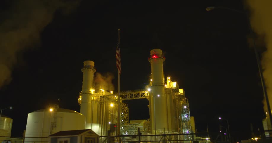 An industrial buidling with lights smoke and chimneys with the sky in the background