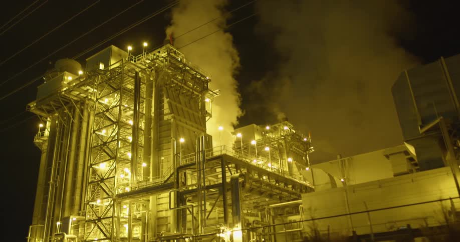 A factory with lights chimneys and smoke with the sky in the background