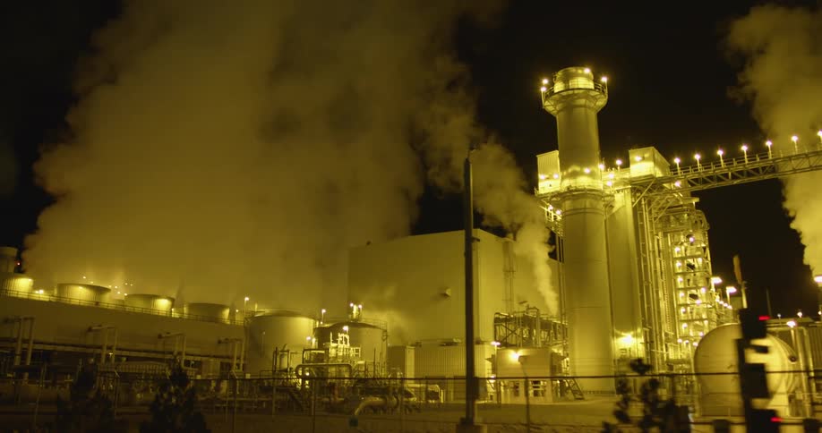 A factory with lights chimneys and smoke with the sky in the background