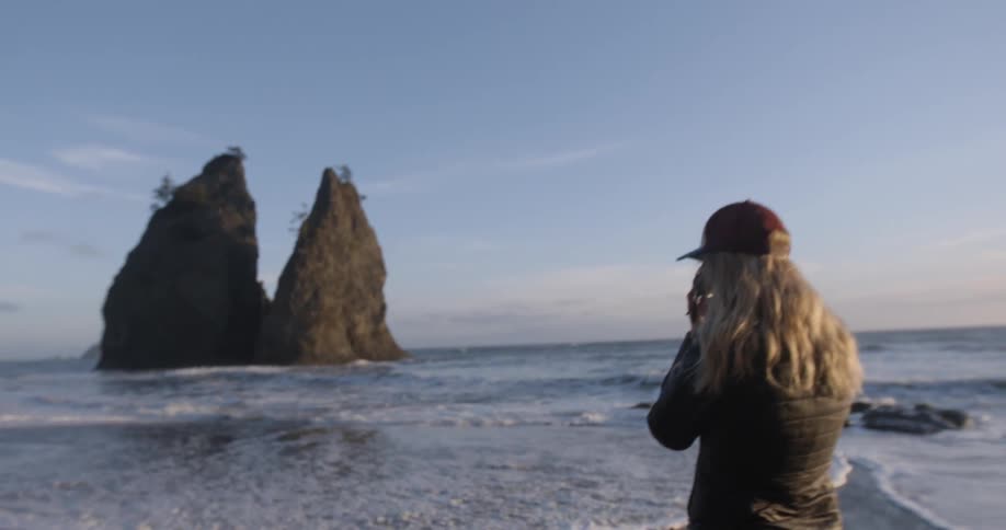Blonde woman wearing a baseball cap taking pictures of large rocks on the beach
