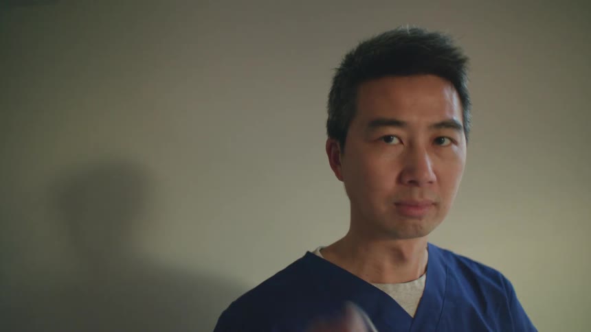 A doctor in blue scrubs removes his surgical mask and smiles while standing against a wall