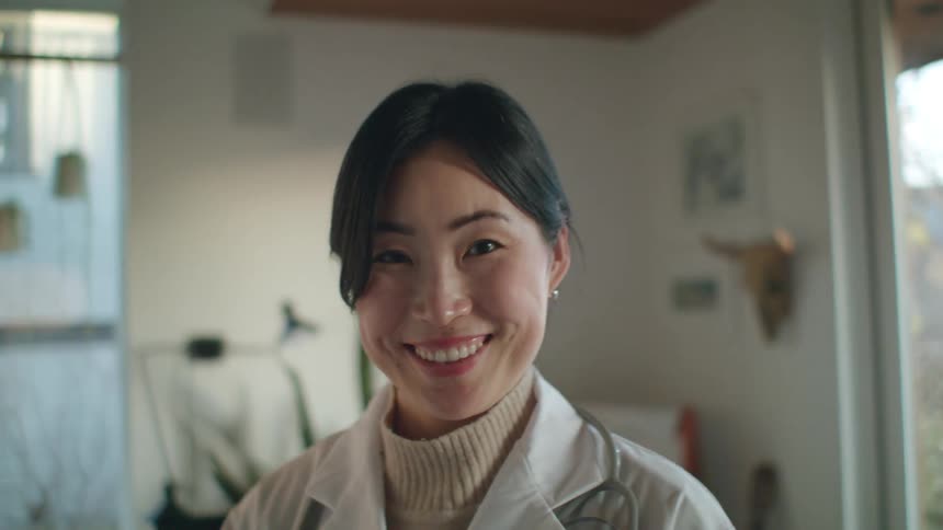 An asian woman wearing a white coat and a stethoscope smiles while standing indoors