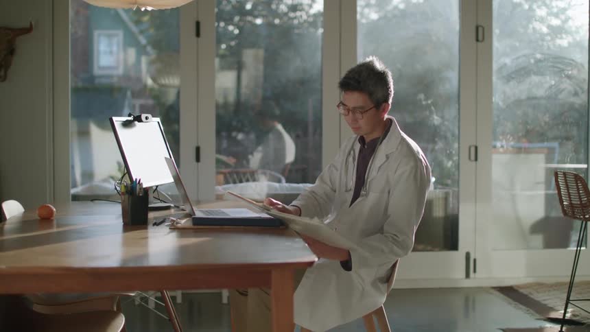 A doctor sits at home and uses his laptop and looks at paperwork