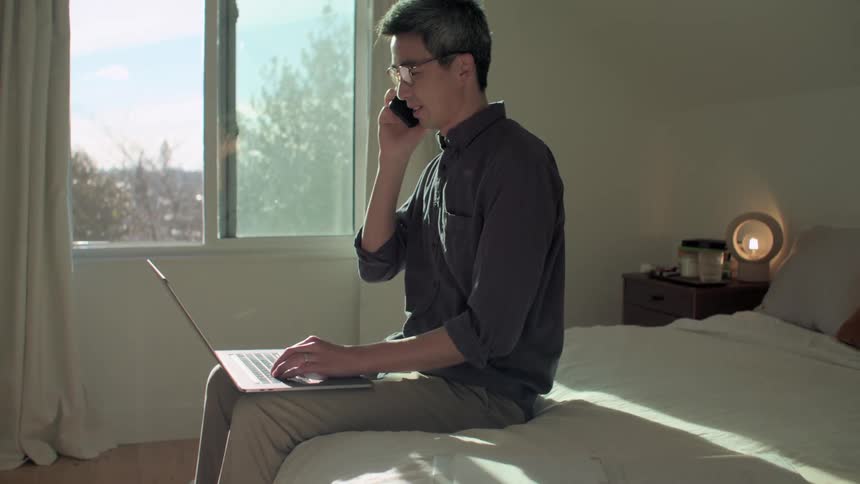 A man is sitting on the edge of a bed with his laptop while talking on a smart phone