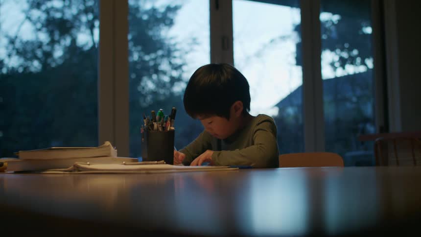 A boy in a room sitting at a table writing on a paper with a pen