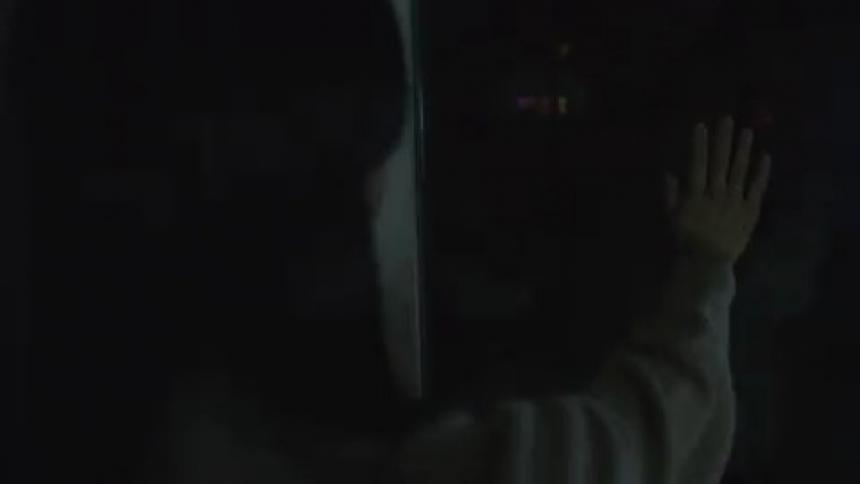 A woman stands with he hand pressed against a window in the dark with flashes of lights
