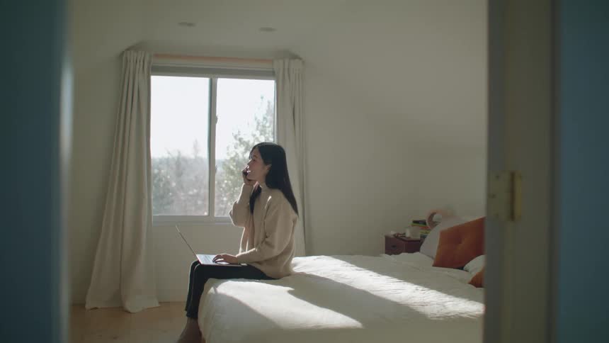 A woman in a bedroom sitting on a bed talking on a smart phone and typing on a laptop