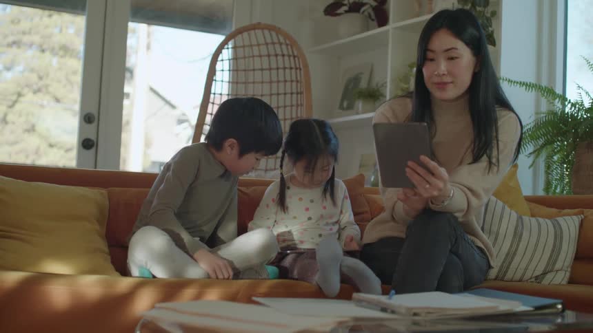 A woman talks via a tablet while her son and daughter play next to her inside on a couch