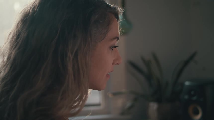A woman sits indoors in a room by a window with a potted plant
