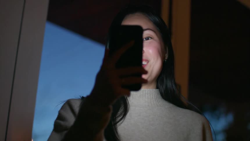 An asian woman holds a phone in her hand as she talks into it while standing indoors at night