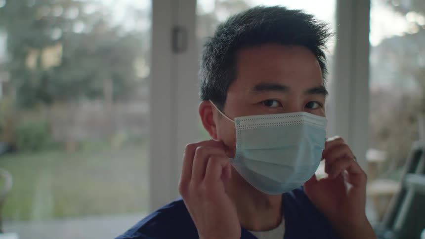 A man in a room with scrubs removing a mask with a door in the background
