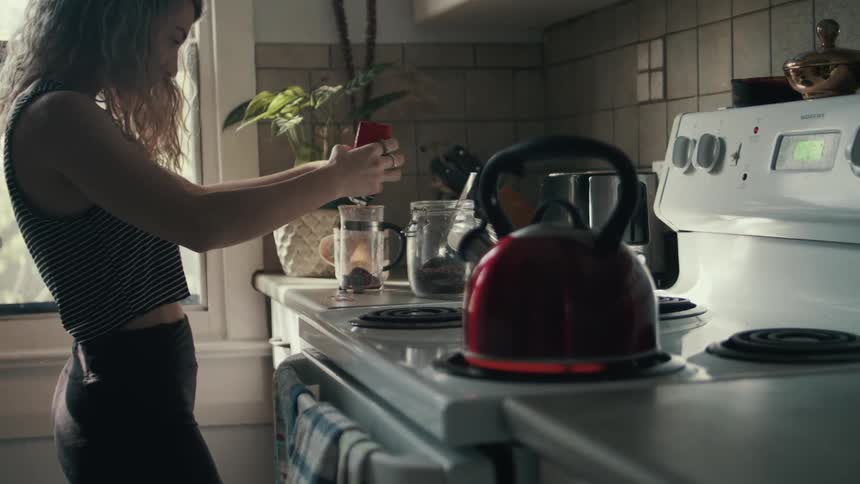 A woman pours coffee grounds into a french press as a kettle heats up on the stove