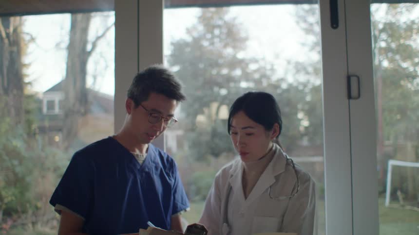 A man with scrubs walking up to a woman with a stethoscope while showing her a clipboard