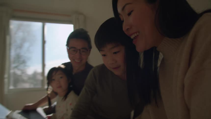 A boy carrying a tablet runs up a staircase and into a bedroom where his parents and sister are waiting on a bed during the day