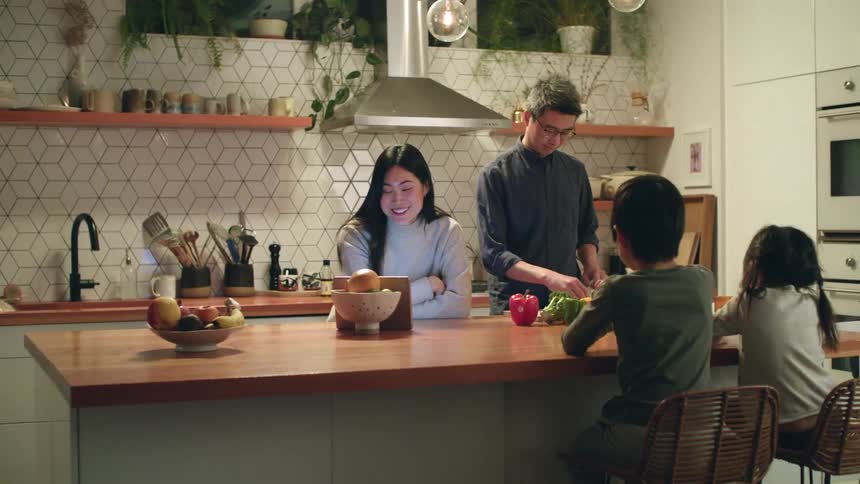 A woman leaning against the kitchen counter while looking at tablet speaks with her husband who is cutting vegetables beside as their children sit and wait on the other side of the counter