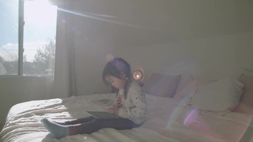 A girl sitting on a bed playing with a tablet