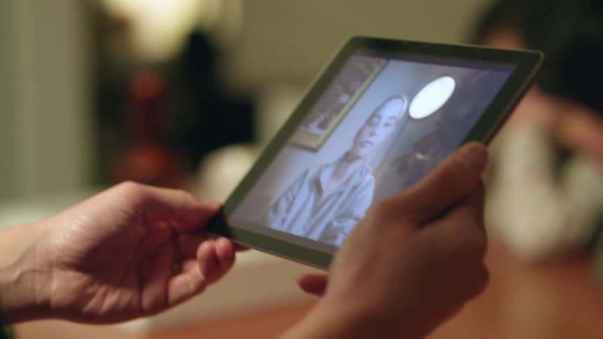 Someone is holding a tablet with a girl on the screen while sitting at a table across from a boy and a girl