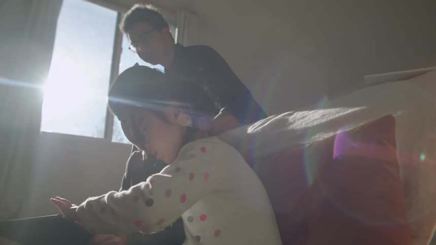 A boy and a girl sitting on the floor next to a bed looking at a tablet with a man with a laptop sitting on the bed