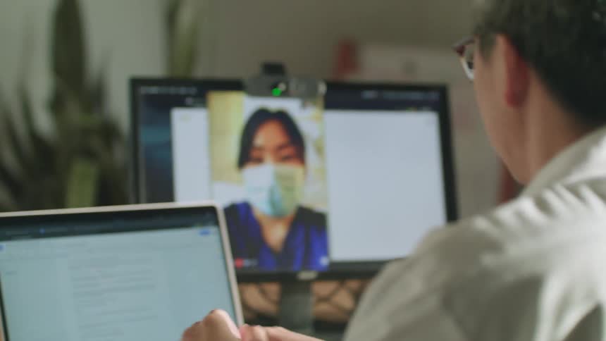 A man participating in a video chat with a masked nurse on a computer lowers the small child sitting in his lap to floor
