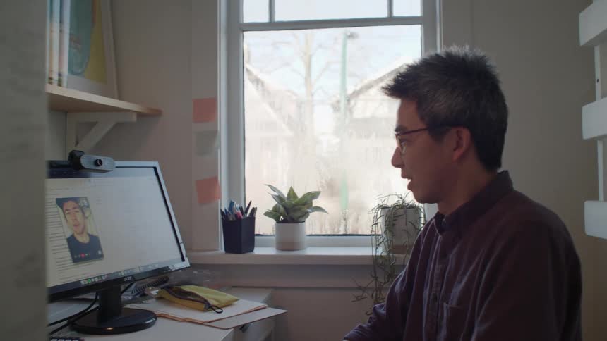 An asian man sits in front of a computer monitor communicating with another male