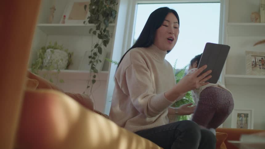 A woman sitting on a couch looking at a tablet with a little girl standing on the couch