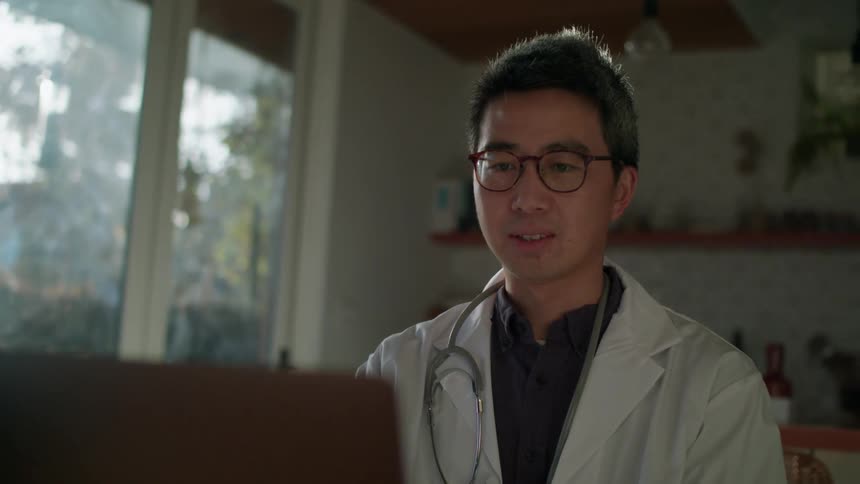 A doctor with a stethoscope and glasses sitting at a table in front of a laptop