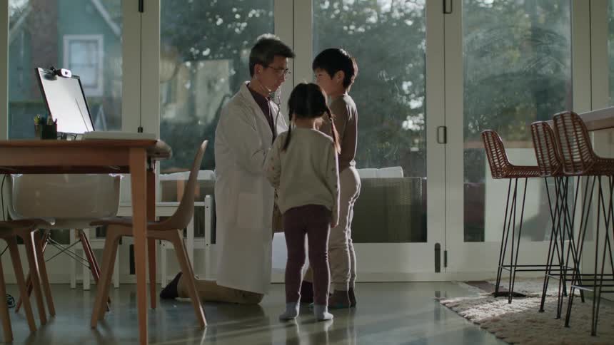A doctor with a stethoscope listening to the heart of a boy with a girl running towards them