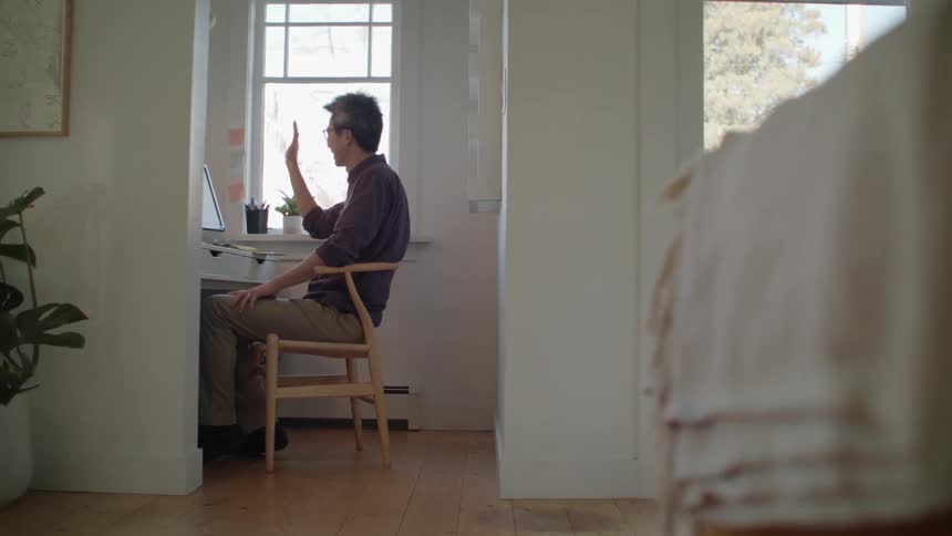 A man sits in a home office waving at his computer monitor
