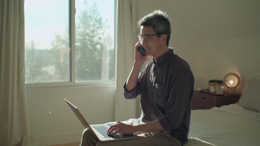 A man sitting on a bed with a laptop answering a phone