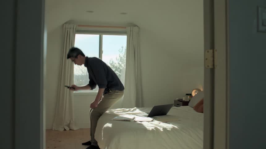 A man in a room talking on a smart phone and sitting on a bed and typing on a laptop
