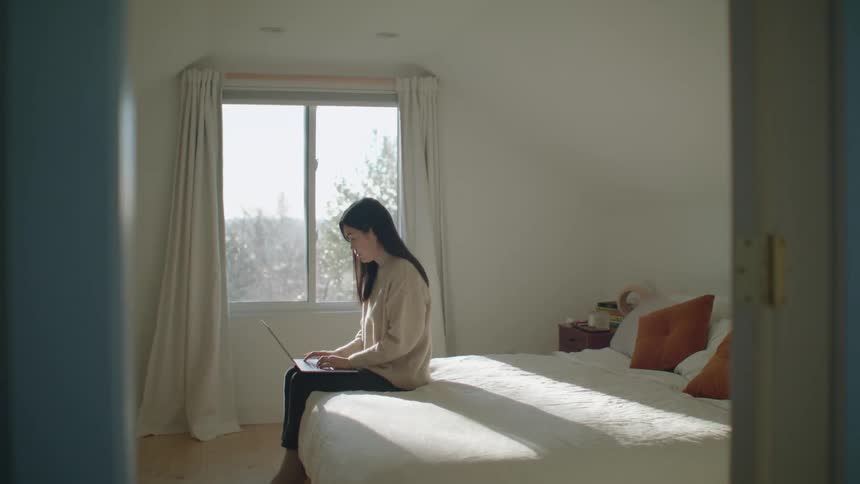 A woman typing on a laptop while sitting on a bed in the daytime