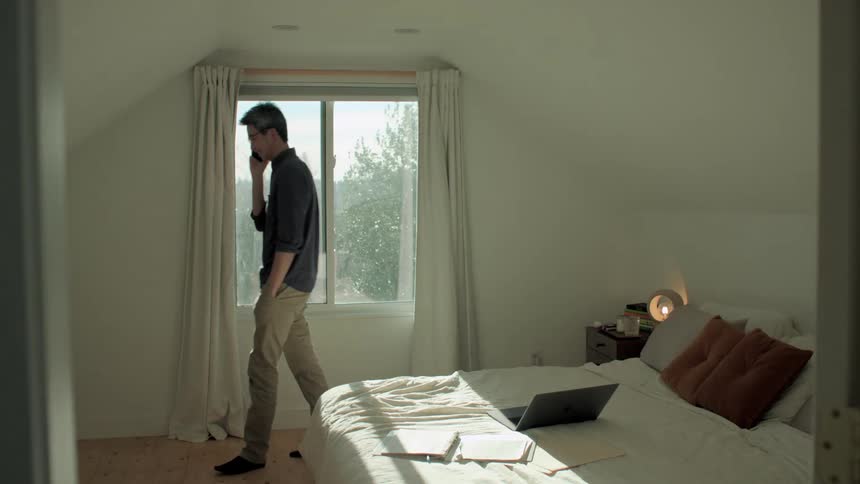 A smiling man paces in front of a window while he talks on a smartphone in a bedroom during the day