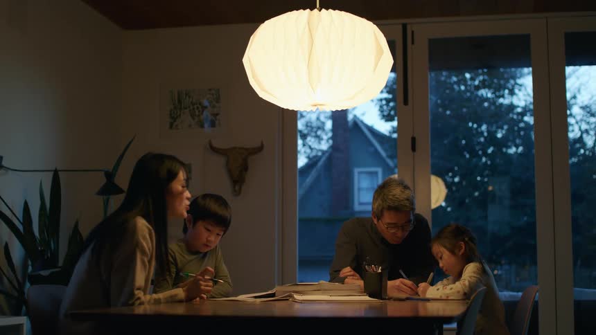 A mother and father help their children with homework as they sit their dining room table in front of a patio door
