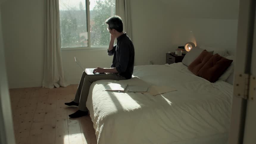 Man sitting in bed at home talks on a smartphone while working on a laptop in the morning