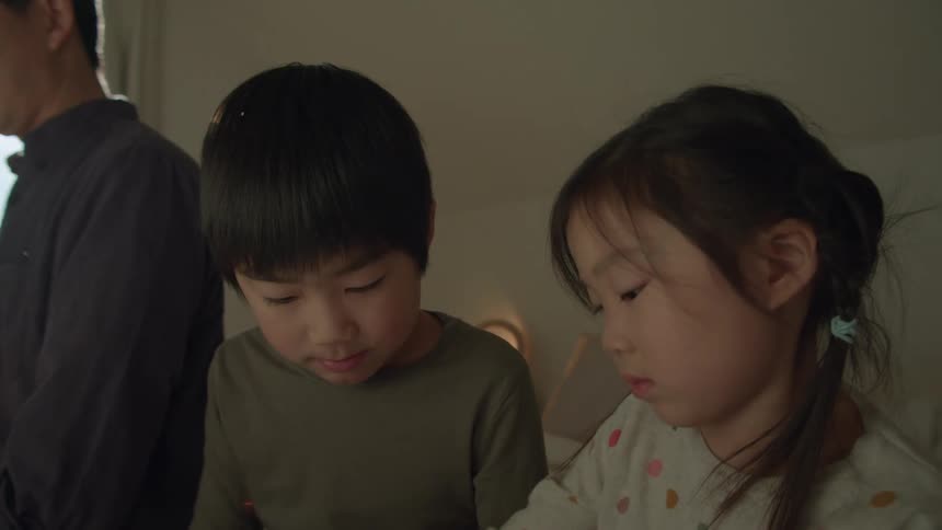 A girl and a boy sitting on a bed looking at a tablet with a man next to them