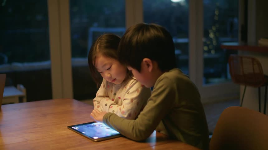A little boy sits next to his sister and they play on a tablet together