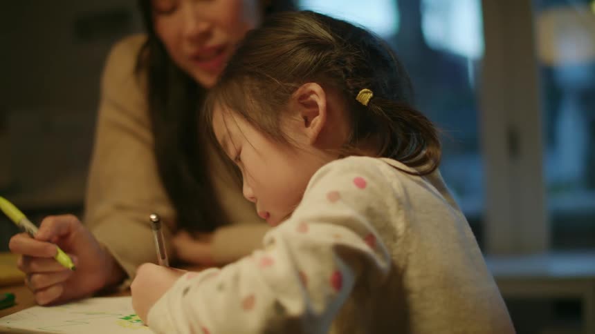 A woman sits at a table at home with her daughter and watches her scribble on paper