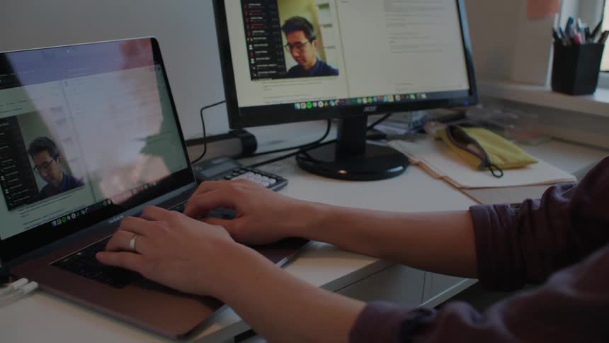 A man sitting at a desk typing on a laptop next to a monitor