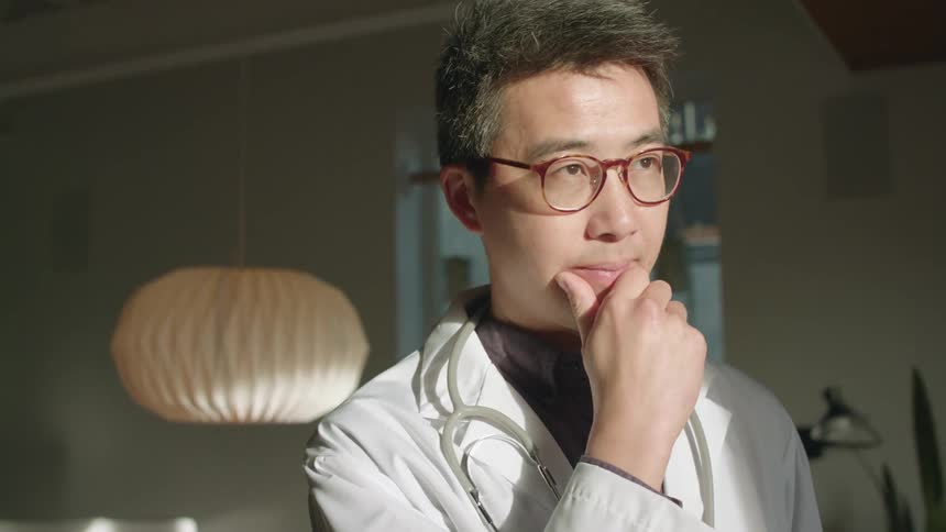 A doctor poses in his white coat and stethoscope while standing in his house thinking with his chin resting on his hand