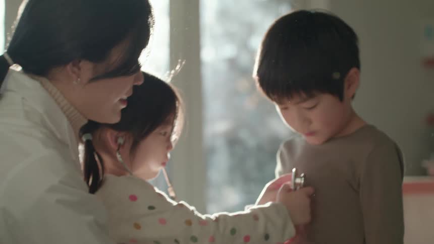 Female doctor teaching her daughter how to use a stethoscope on his brother at home