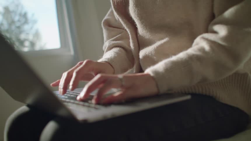 A woman finishes a call on her smartphone and begins typing on a laptop as she sits on a bed in a bedroom during the day