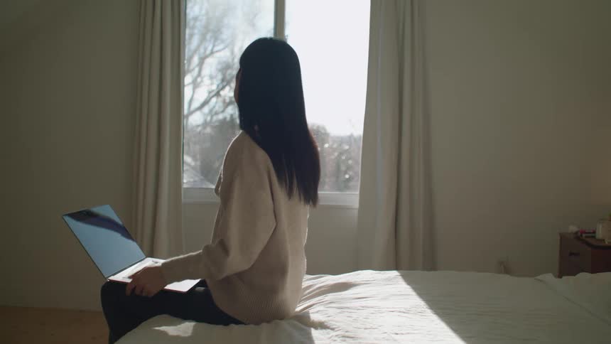 A woman sitting on a bed talking on a smart phone using a laptop and looking out of the window