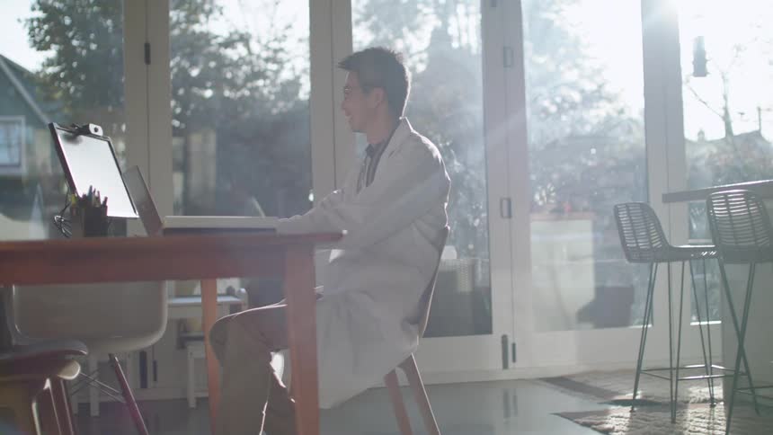 A doctor sits at home and waves as he video chats on a laptop