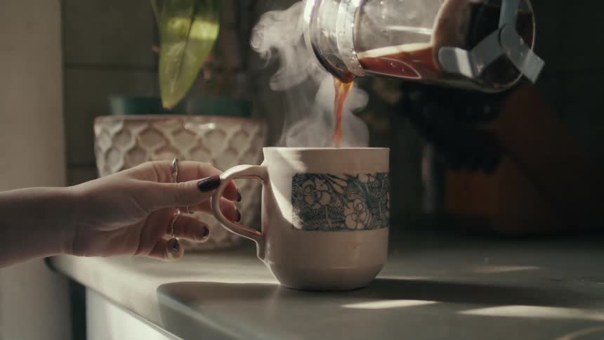 A woman pouring coffee into a cup on a counter with a plant in the background