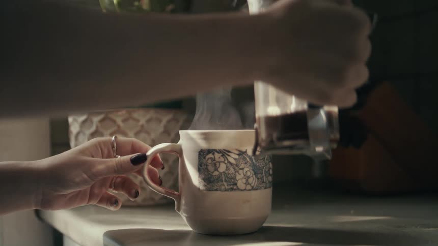 A woman pours steaming coffee from a small coffee pot into a coffee mug