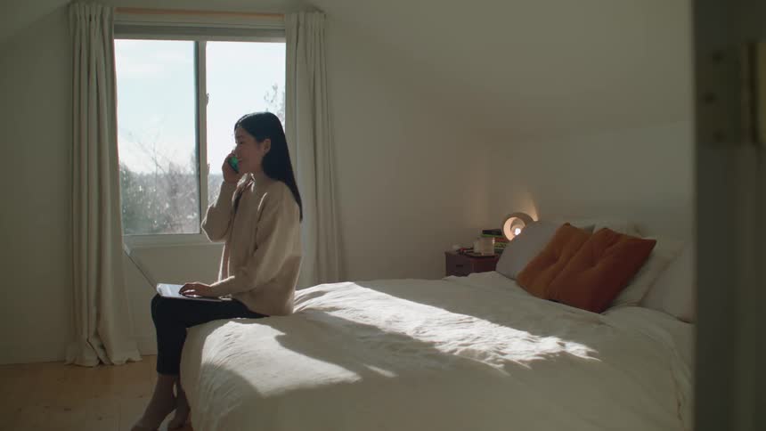 A woman sitting on a bed typing on a laptop and talking on a smart phone