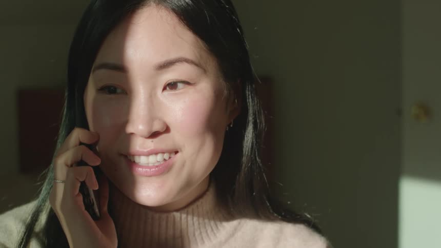 An asian woman smiles and talks on a cell phone while standing indoors during the day