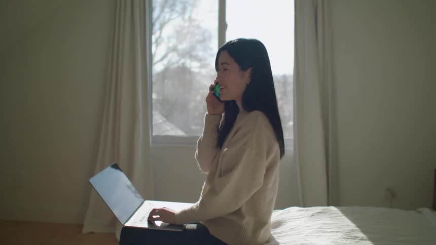 An asian woman sits on a bed typing on a laptop and answering a cell phone